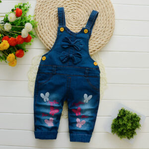 Salopette en jean motif papillon pour fille couleur bleu denim. Les papillon sont sur les jambes, de couleur rose et blanche. La salopette est posée sur un rond en corde avec des fleurs sur le côté.