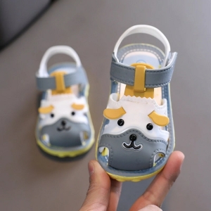 Sandale pour bébé fille de couleur grise et de forme de petit chien tenu par une main et l'autre posé sur un canapé gris
