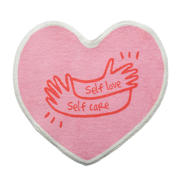 Tapis de chambre pour fille en forme de cœur de couleur rose. Sur le dessus un imprimé de deux mains avec l'inscription self love self care de couleur rouge.