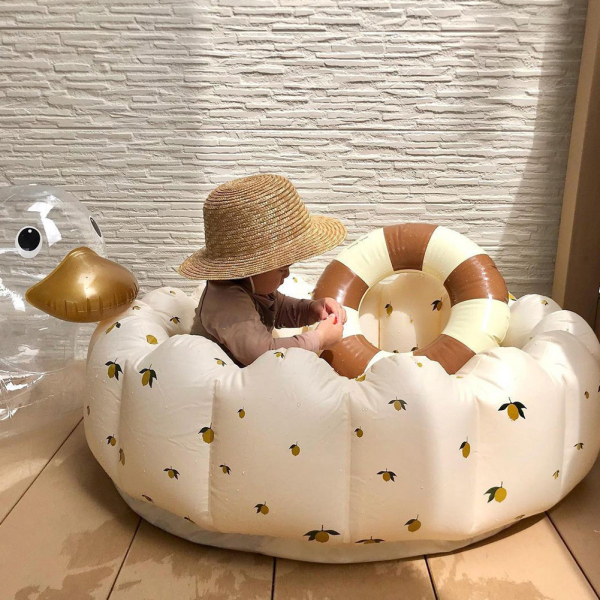 Piscine gonflable pour enfant de couleur blanche avec des motifs d'olivier, posée sur un sol en bois devant un mur blanc avec un bébé avec un chapeau de paille à l'intérieur