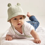 Bébé à plat ventre sur un tissu blanc nid d'abeille portant un body blanc, un jean et un chapeau en molleton beige avec petites oreilles et cordelette.