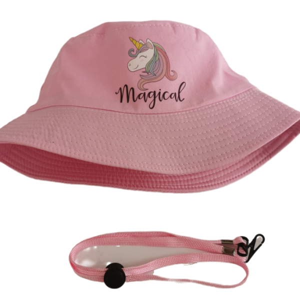 Sur fond blanc, chapeau de type bob rose clair avec une illustration de licorne blanche à la crinière multicolore et à la corne dorée et inscription Magical sur l'avant et cordelette de maintien posée à côté avec bouton de serrage en plastique noir et crochets en plastique noir.