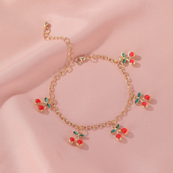 Le bracelet en maille avec breloque cerise pour bébé fille est posé sur un drap rose, fait en maillon de chaine avec cinq petits pendentifs en cerise. Il possède un fermoir ajustable