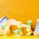 Figurines Smiski Dressing, présentant des figurines phosphorescentes en train d'essayer des vêtements, sur un fond jaune vif.