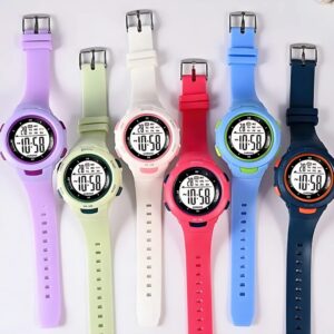 Six montres digitales multifonctions pour filles, disposées côte à côte. De gauche à droite, les couleurs sont : violet, vert clair, blanc, rose, bleu et bleu marine. Chaque montre possède un cadran rond avec un affichage digital, des boutons sur les côtés et un bracelet en silicone assorti à la couleur du boîtier. Les montres affichent l'heure, la date, le jour de la semaine et disposent de fonctions chronomètre et alarme.