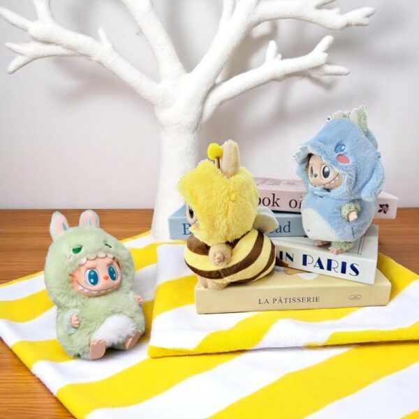 3 peluches labubu portant des costumes d'animaux en peluche, disposés sur une serviette jaune et blanche, sur une table en bois avec un arbre blanc en fond
