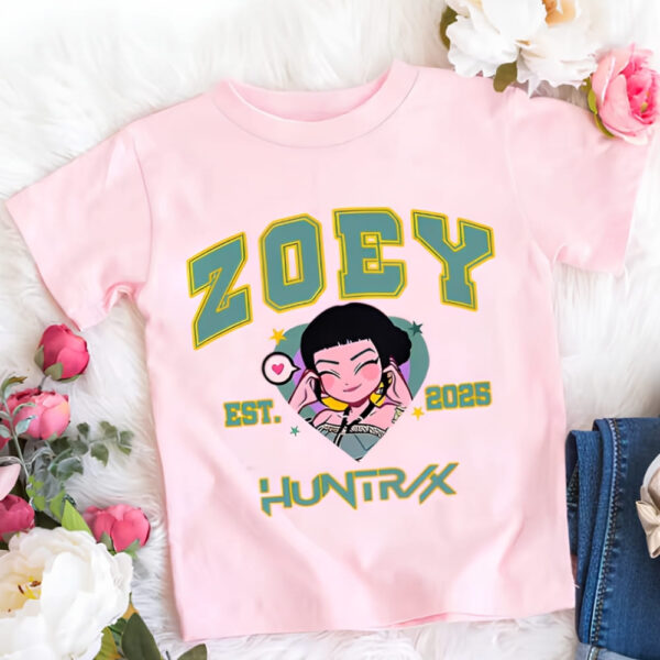 T-shirt fille K-pop Demon Hunters rose du personnage Zoey. Son prénom est écrit en majuscules vertes ainsi que Huntrix sous le portrait de Zoey en version cartoon. Le t-shirt est posé à côté de fleurs.