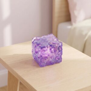 Un cube squishy anti-stress Needoh à paillettes violet posé sur une petite table en bois, dans une pièce douce et lumineuse.